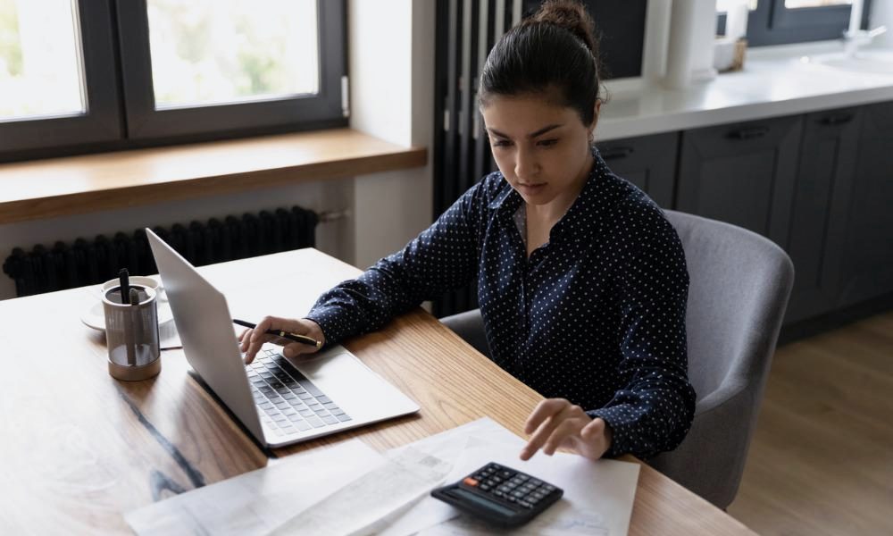 A woman works through paperwork with a laptop and calculator at a desk.